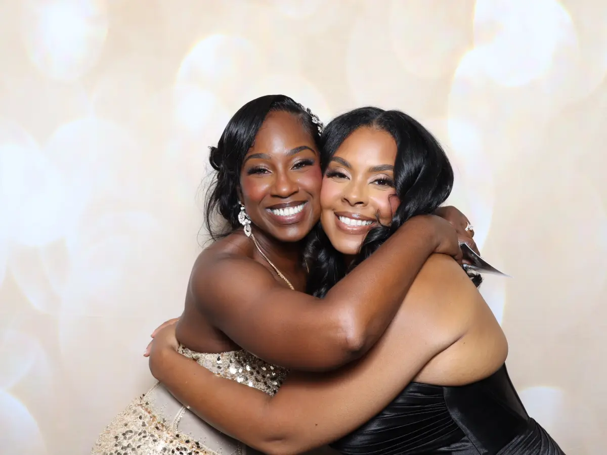 Two guests in formal gowns hugging in front of a champagne bokeh photo booth backdrop at Bella Blanca Event Center