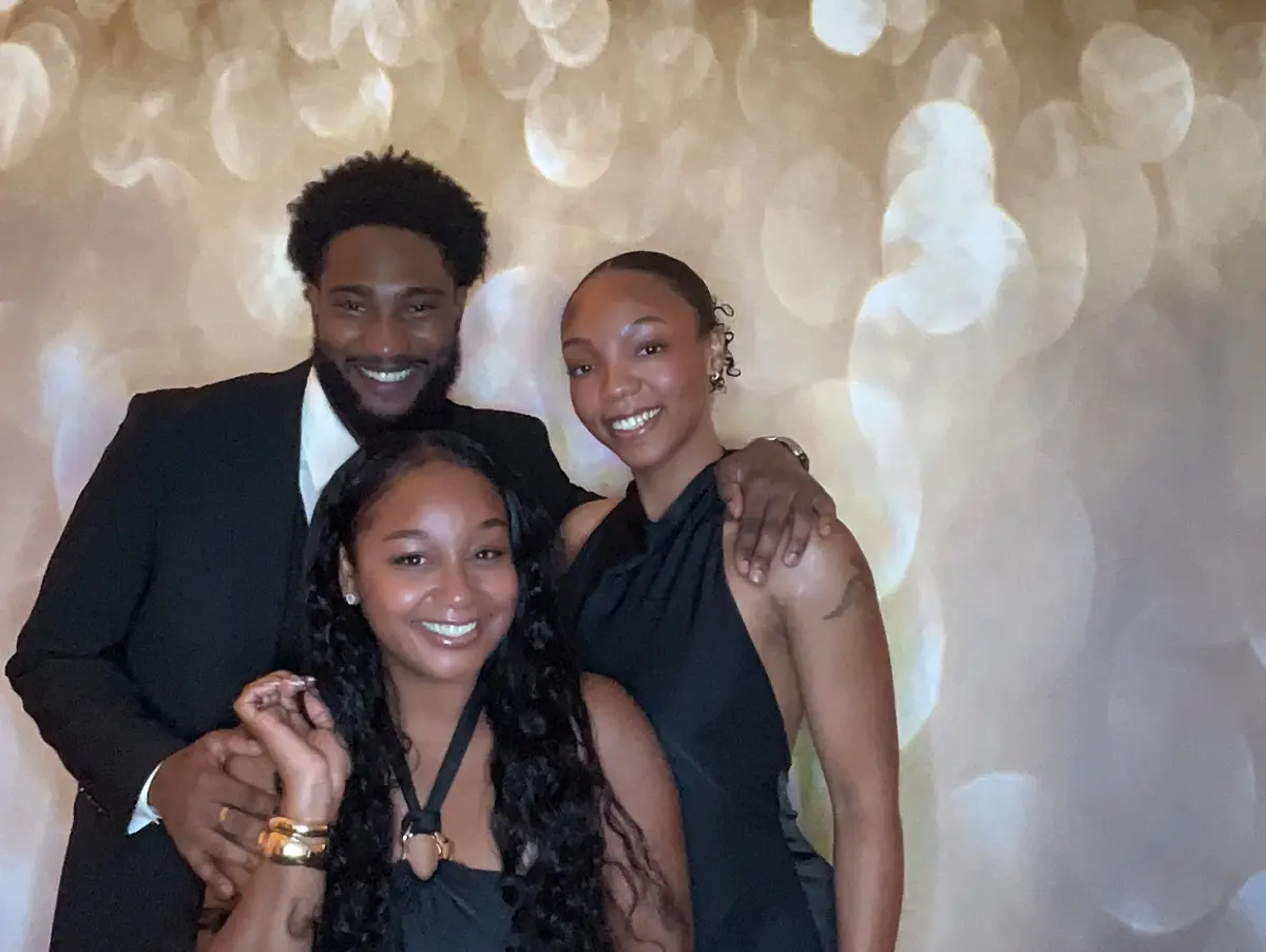 Three friends in black formal wear smiling in front of a gold glitter bokeh backdrop