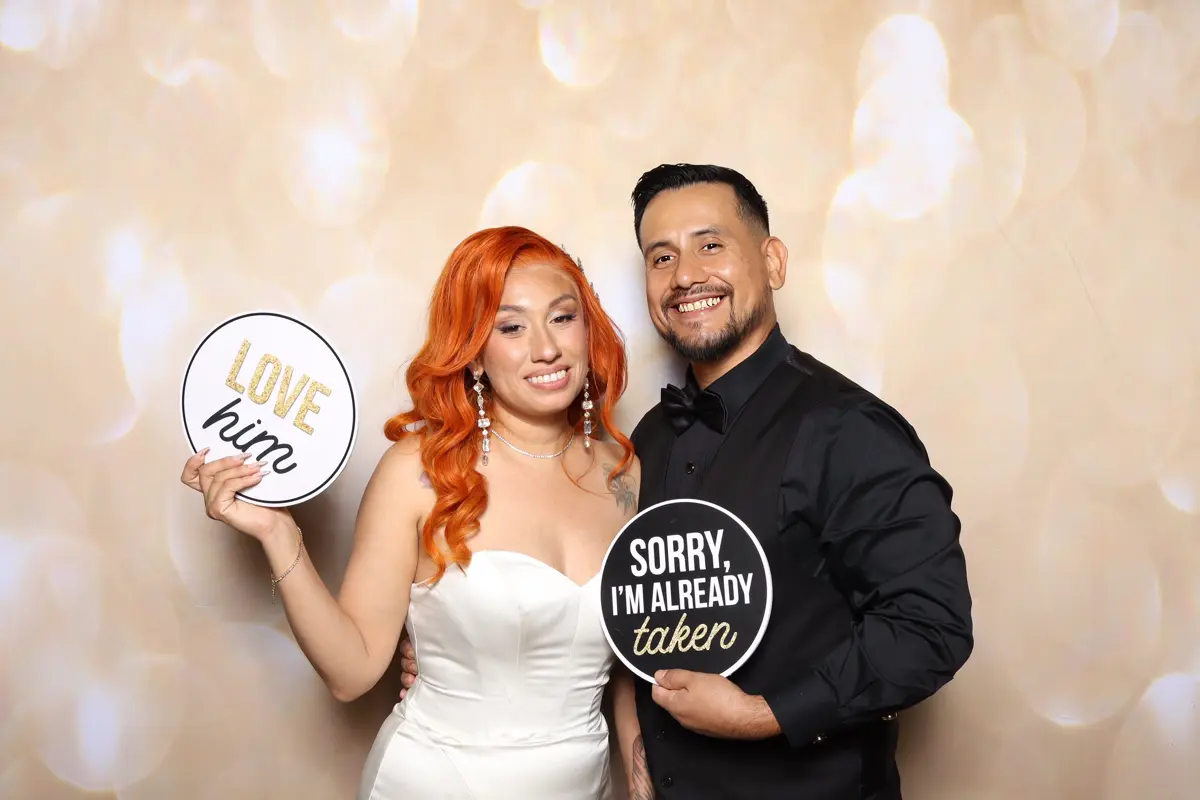 Bride Aley holding a Love Him sign and groom Julian holding a Sorry I’m Already Taken sign at their wedding photo booth at Hart & Main in Santa Clarita