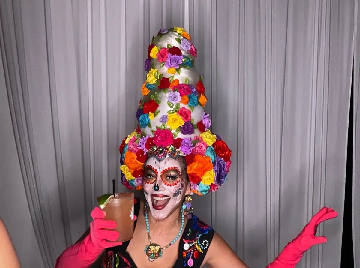 Guest in a tall rainbow rose headdress and sugar skull makeup raises a cocktail at the Catrina Ball photo booth