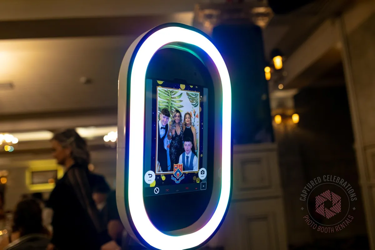 A photo booth at an event displays a group photo of four smiling people in formal attire. The booth has a colorful neon rim.