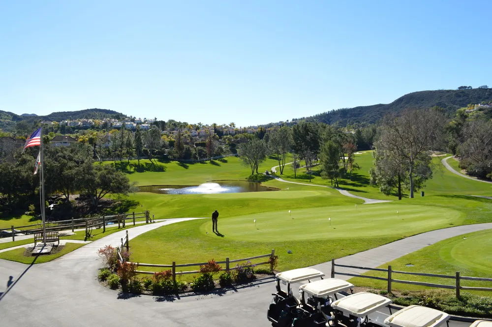 Golf course with a person putting on a green. Golf carts in the foreground, pond and flagpole nearby, surrounded by hills under a clear sky.