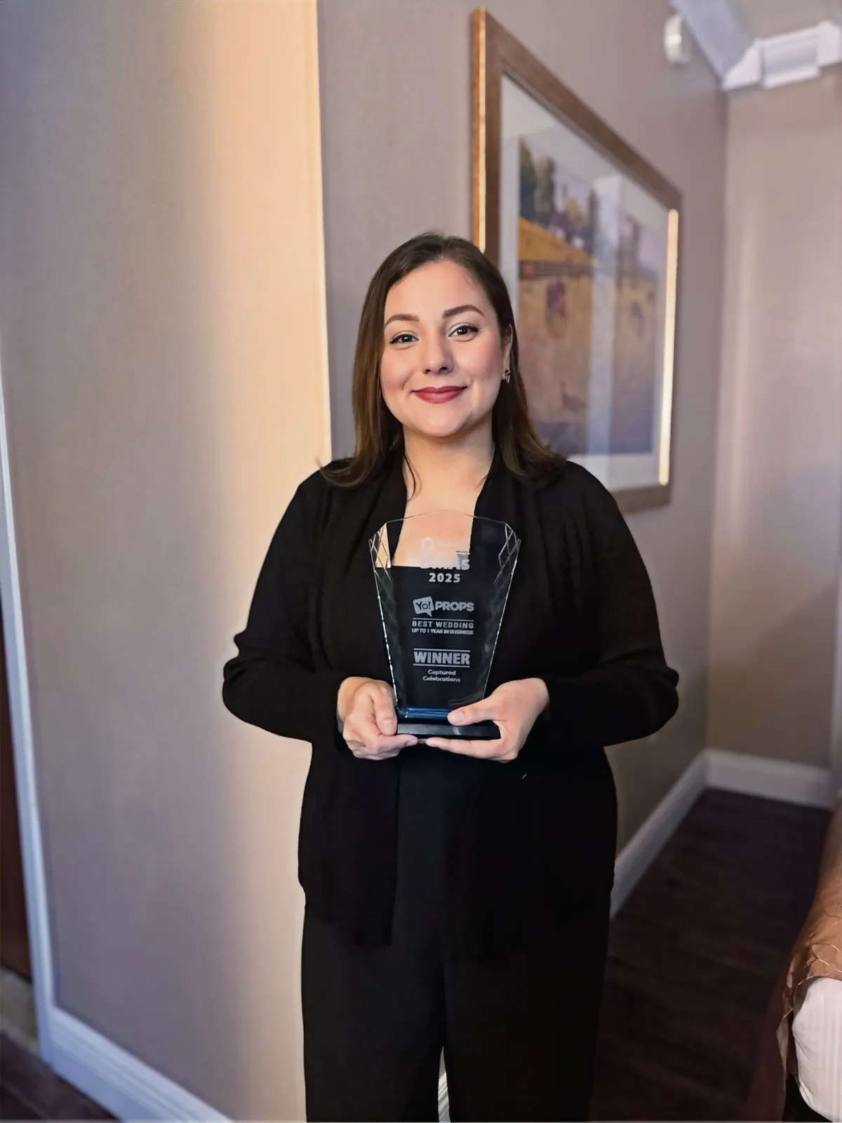 Liz smiling and holding a "Winner" trophy in a warmly lit room with a framed picture on the wall. She's wearing a black outfit.