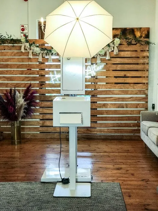 White photo booth with large umbrella in front of a wooden backdrop. Neon hashtag sign partially visible. Decorative plants nearby. Cozy setting.