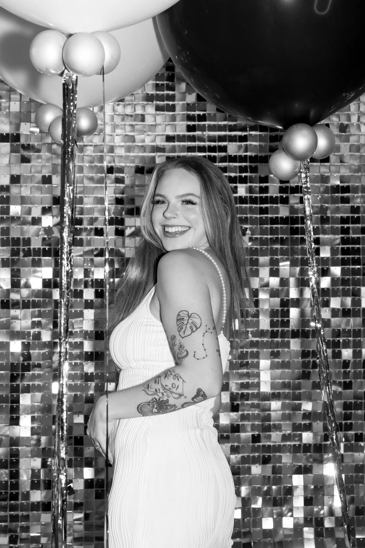 Smiling woman in a white dress with tattoos stands in front of a shimmering mosaic wall, holding balloons. Festive and joyful atmosphere.