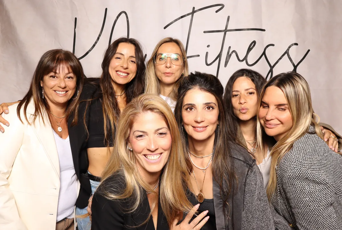 Seven women smiling and posing in front of a "K+Fitness" backdrop. Casual attire, joyful mood, diverse hair colors.