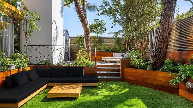Modern patio with black cushions on wooden seating, a wooden coffee table, green grass, and lush plants. Bright and serene atmosphere.
