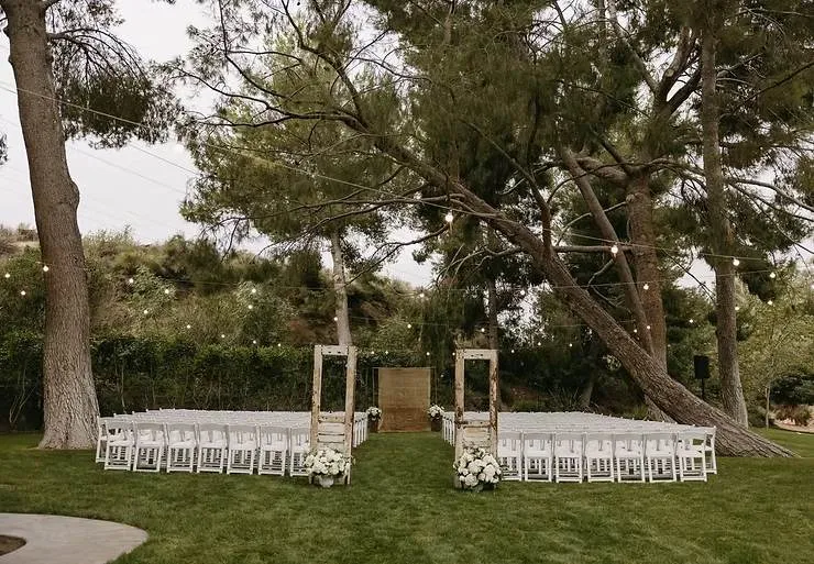 Outdoor wedding setup with white chairs on green grass, surrounded by trees. String lights hang above; rustic wooden door frames and flowers line the aisle.