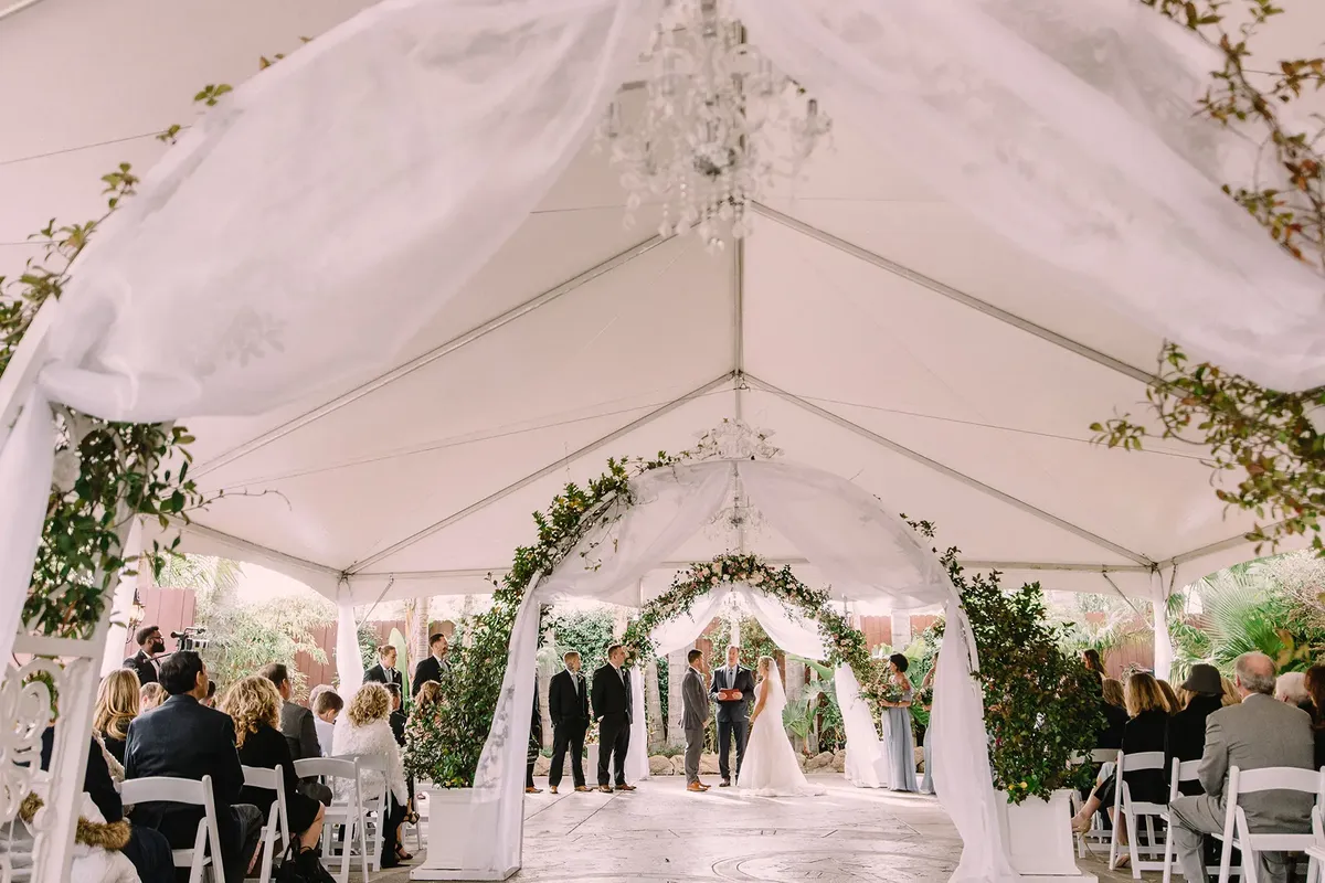 Wedding ceremony under a decorated tent with greenery and draped fabric. Bride and groom stand together, surrounded by guests in formal attire.