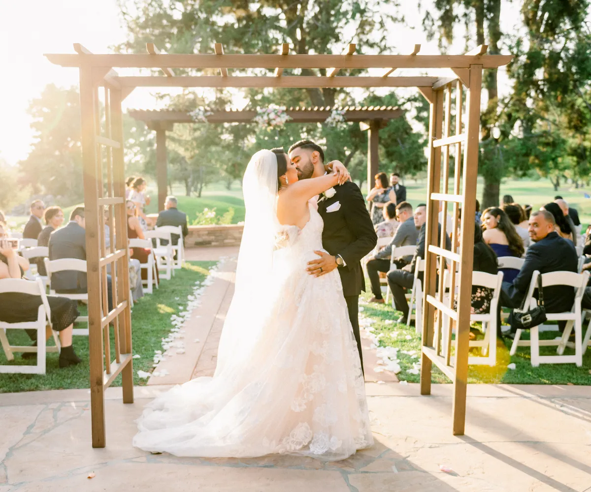 Bride and groom kiss under a wooden arch at an outdoor wedding, surrounded by seated guests. Sunlight filters through trees, creating a warm glow.