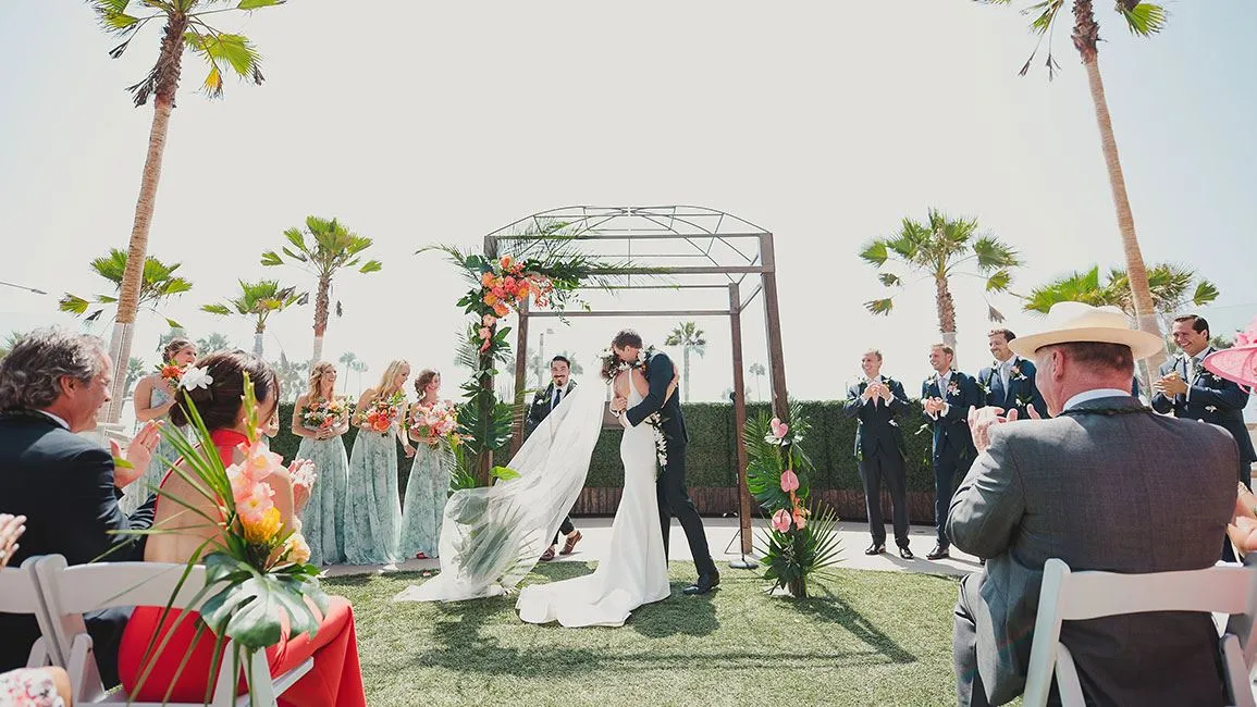 Bride and groom kiss under floral arch with joyful guests clapping. Sunny outdoor wedding with palm trees and vibrant bouquets.