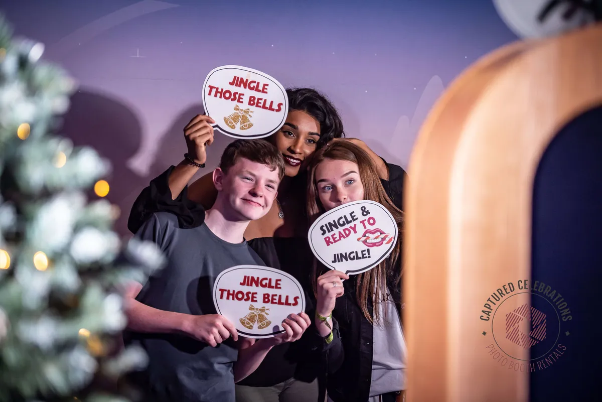 Three people pose happily with festive signs reading "Jingle Those Bells" and "Single & Ready to Jingle" in a decorated setting.