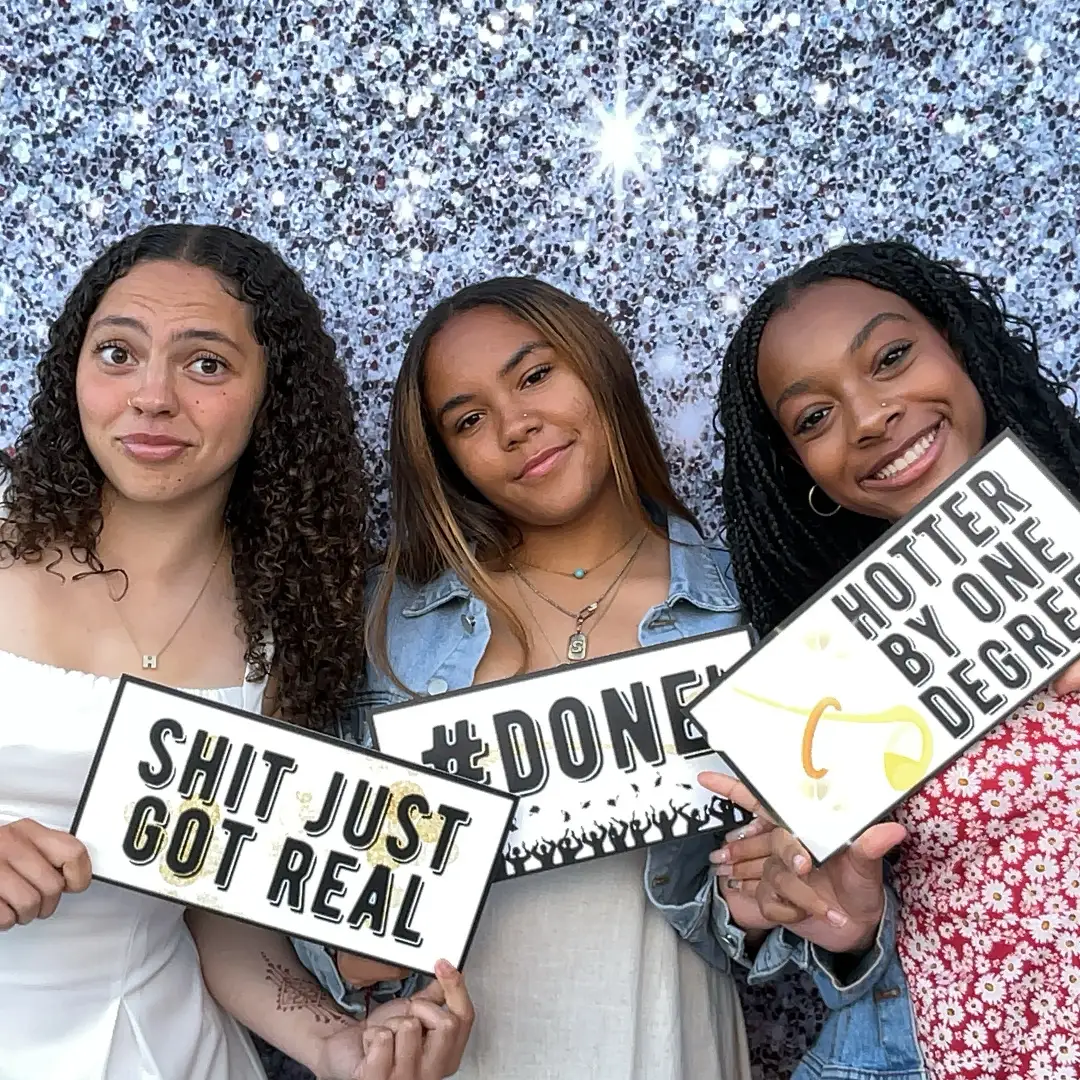 Three women, graduation photo booth props: "Shit just got real", "#Done", "Hotter by one degree"