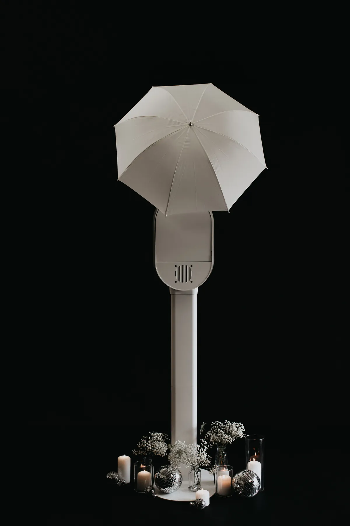 White umbrella on a stand, surrounded by candles, flowers, and disco balls on a black background, creating a serene and elegant scene.